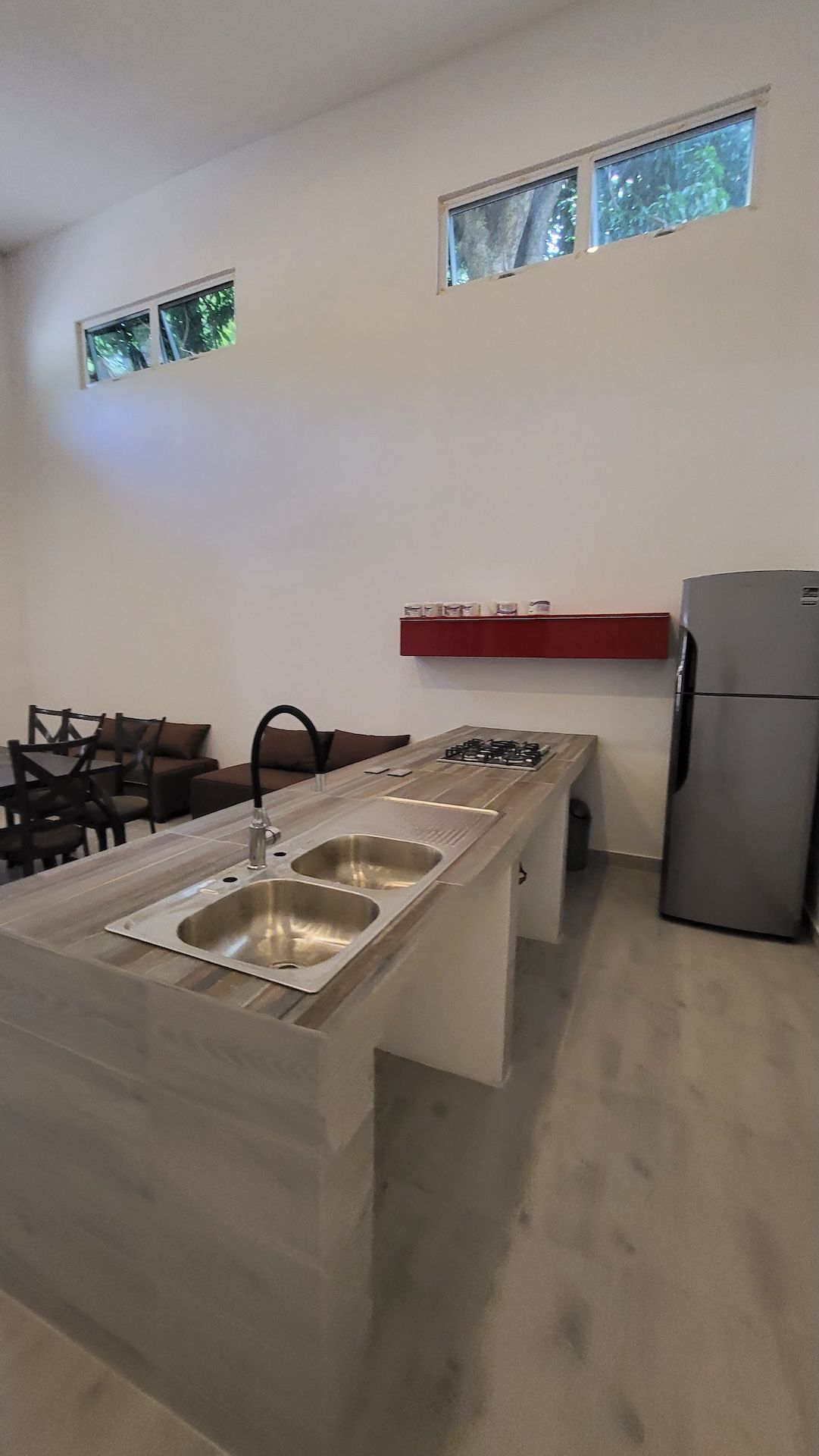 A compact kitchen with a central island, a built-in sink, a gas stove, and a refrigerator. There are two windows on the wall, and a red shelf above the sink.