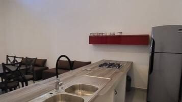 A compact kitchen with a central island, a built-in sink, a gas stove, and a refrigerator. There are two windows on the wall, and a red shelf above the sink.