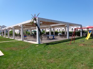 A covered pavilion with tables and chairs, a playground with a slide, and a grassy area.