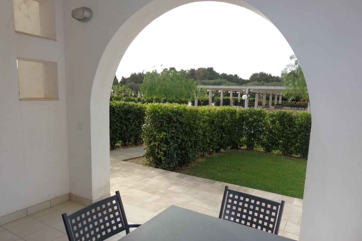A patio area with a table and chairs, a white wall, and a view of a garden and a pergola.