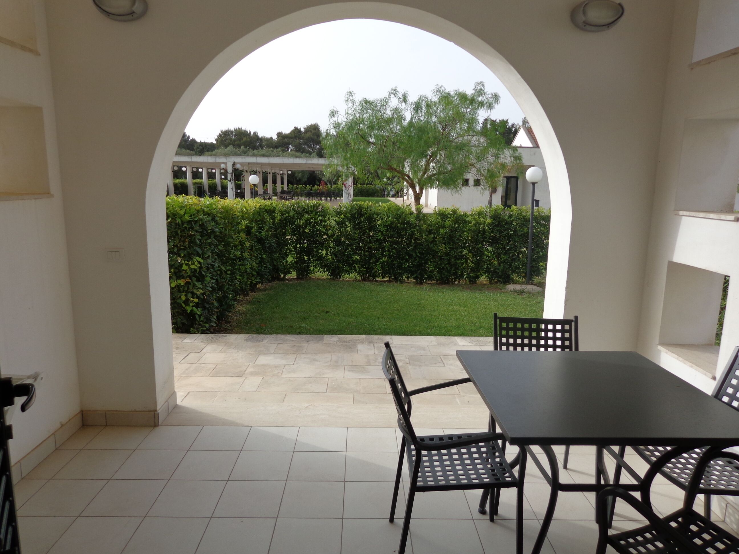 An outdoor patio area with a table and chairs, a tiled floor, and a view of a well-maintained garden through an archway.
