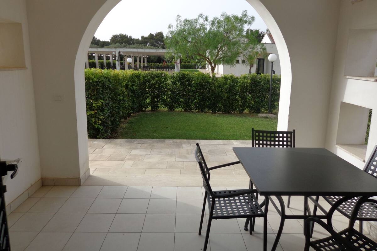 An outdoor patio area with a table and chairs, a tiled floor, and a view of a well-maintained garden through an archway.