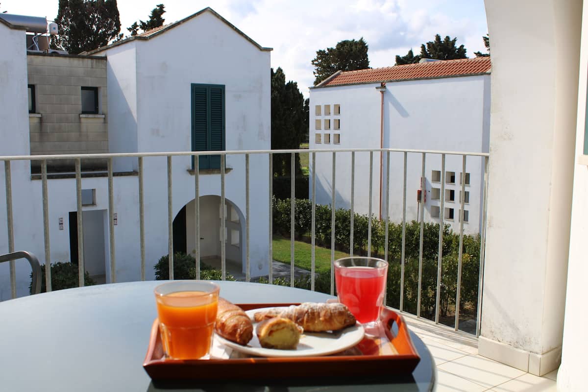 A balcony with a table set for breakfast, including croissants and juice, overlooking a courtyard with buildings and greenery.