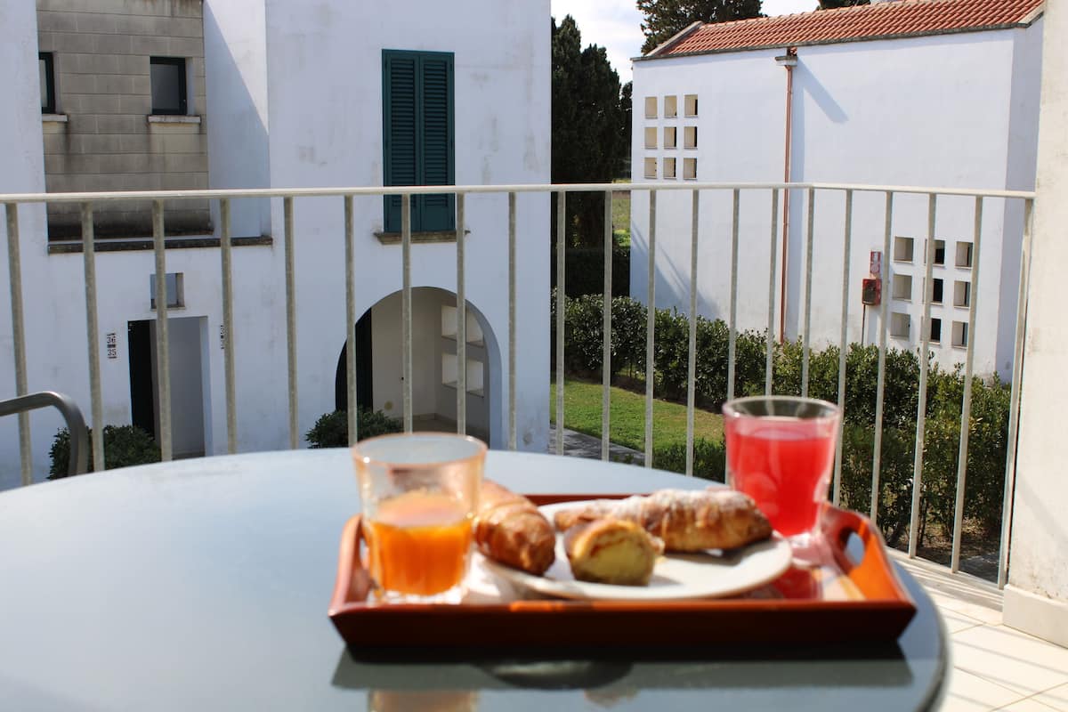A balcony with a table set for breakfast, including croissants and juice, overlooking a white building with a tiled roof.