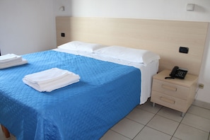 A neatly made bed with a blue bedspread and white pillows, a bedside table with a phone, and a folded white towel on the bed. - Residence Alimini Blu (Otranto)