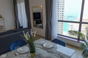 A modern living room with a sofa, a dining table set with plates and cutlery, a television, and a large window with a view of a cityscape and a body of water. - JBR Rimal Apartments (Dubai)