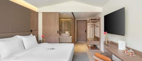 A hotel room with a large bed, a desk with a chair, a television, a mirror, and a bathroom visible through an open door.