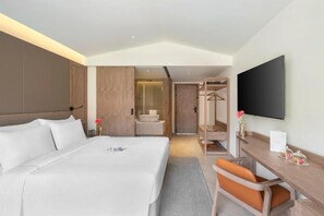 A hotel room with a large bed, a desk with a chair, a television, a mirror, and a bathroom visible through an open door. - Zana in The Wild, Dhikuli (Ramnagar)