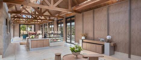 A spacious interior with a high ceiling, wooden beams, a central seating area with a round table, and a view through large windows.