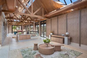 A spacious interior with a high ceiling, wooden beams, a central seating area with a round table, and a view through large windows.
