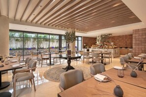 A spacious dining area with multiple tables and chairs, a central decorative plant, and a view of greenery outside.