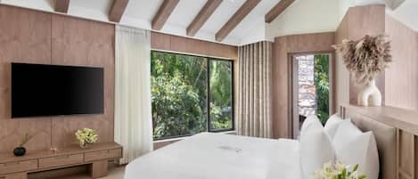 A modern bedroom with a large bed, a flat-screen TV, a wooden bench, and a view of greenery outside.