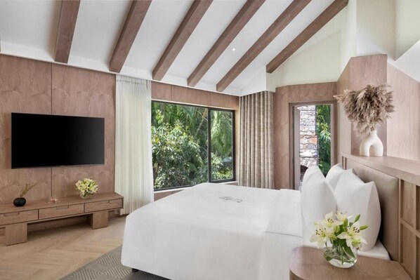 A modern bedroom with a large bed, a flat-screen TV, a wooden bench, and a view of greenery outside.