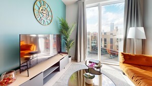 A modern living room with a flat-screen TV, a sofa, a coffee table, a decorative clock, and a large window with a city view. - Charles Hope Apartments - Leeds (Leeds)