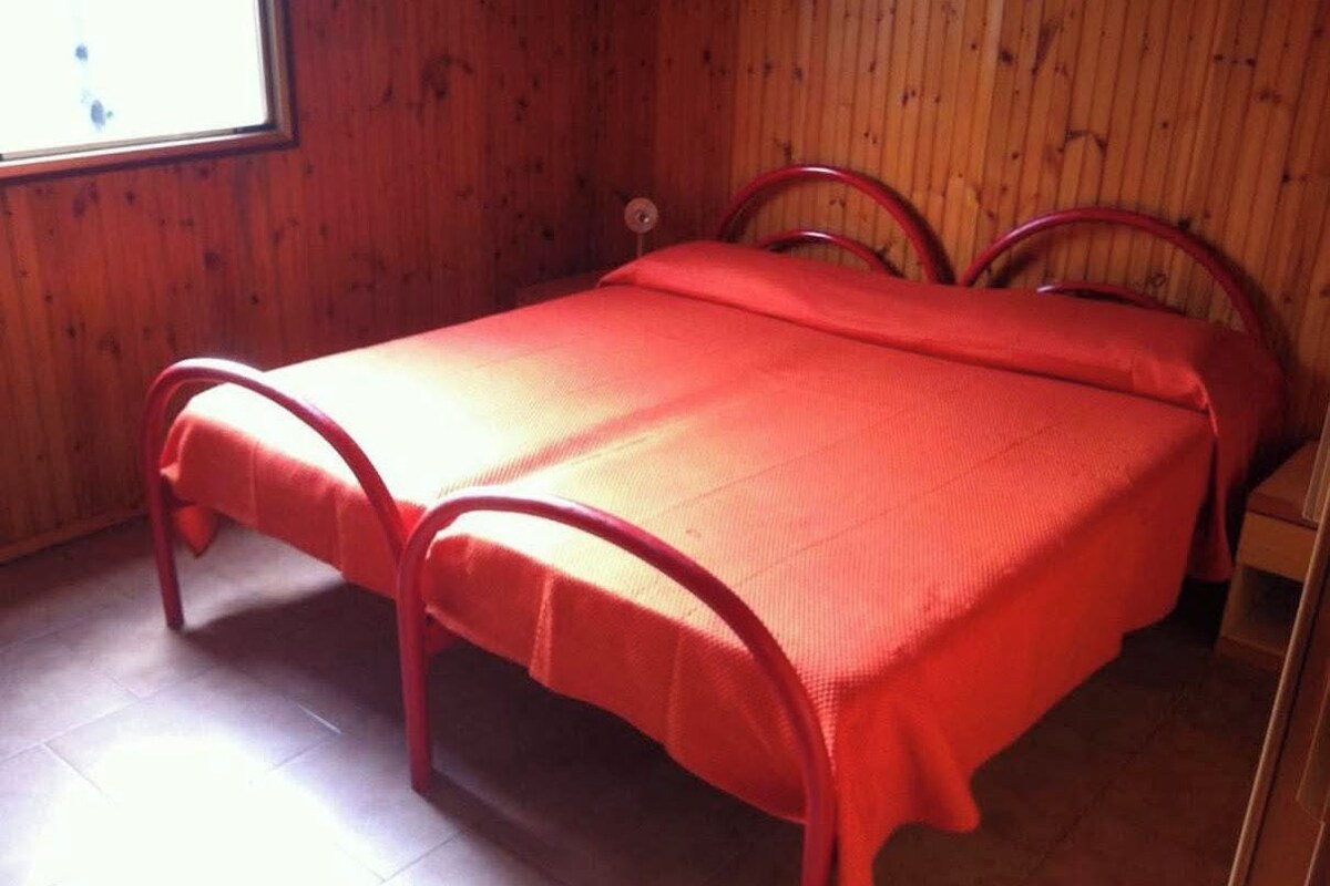 Una cama individual con una colcha roja en una habitación con paredes de madera y un ventanal.