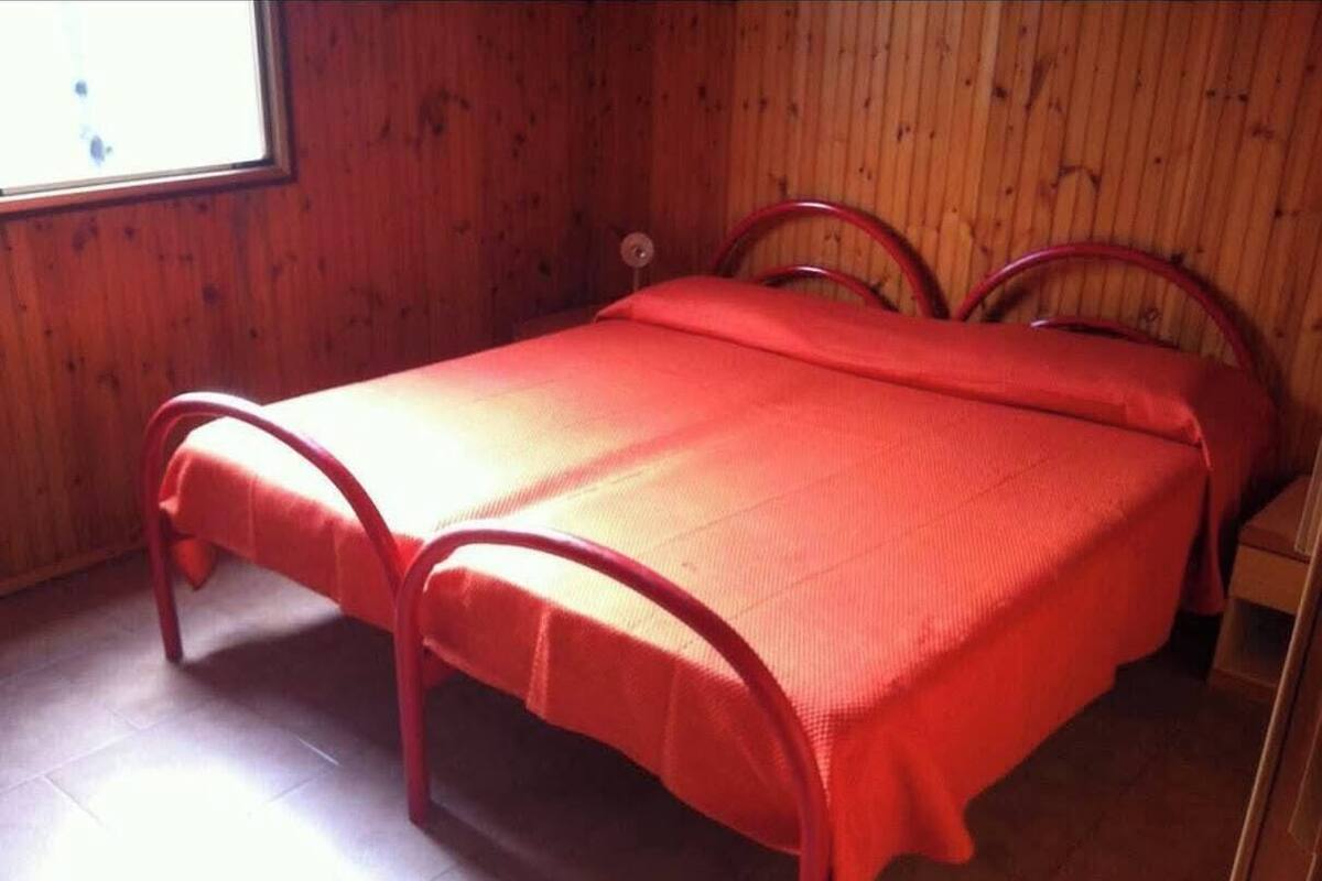 Una cama individual con una colcha roja en una habitación con paredes de madera y un ventanal.
