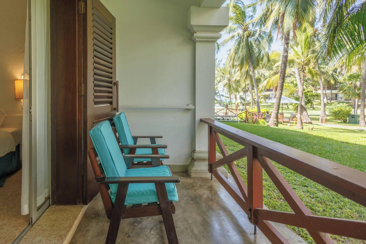 Een balkon met een houten hekwerk, een turquoise stoel en uitzicht op een gazon met palmbomen.
