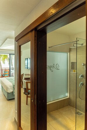 A hotel room with a glass shower enclosure, a bed with a white cover, a desk with a chair, and a view of the beach.