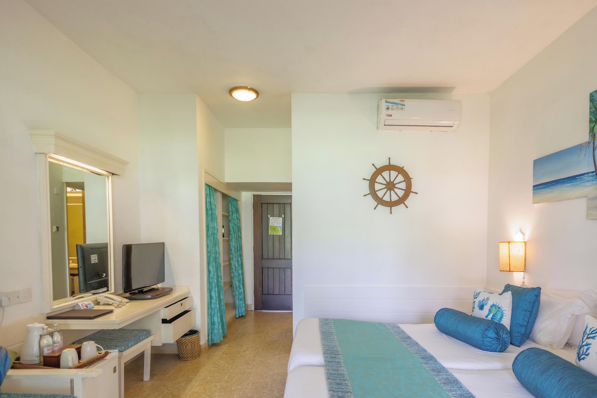 A hotel room with a bed, a desk with a computer, a TV, a wall-mounted air conditioner, a wall clock, and a painting of palm trees.