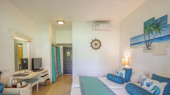 A hotel room with a bed, a desk with a computer, a TV, a wall-mounted air conditioner, a wall clock, and a painting of palm trees. - Heritage Hotels Ltd (Mombasa)