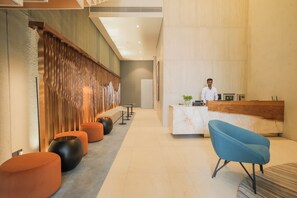 A modern reception area with a wooden accent wall, a reception desk, a blue chair, and a seating area with orange and black round ottomans. - Sesaya Poshtel & Bistro (Sri Jayawardenepura Kotte)