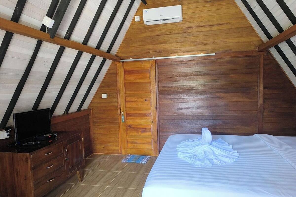 A room with a sloped ceiling, wooden walls, a bed with a folded towel, a TV on a wooden cabinet, and an air conditioning unit.