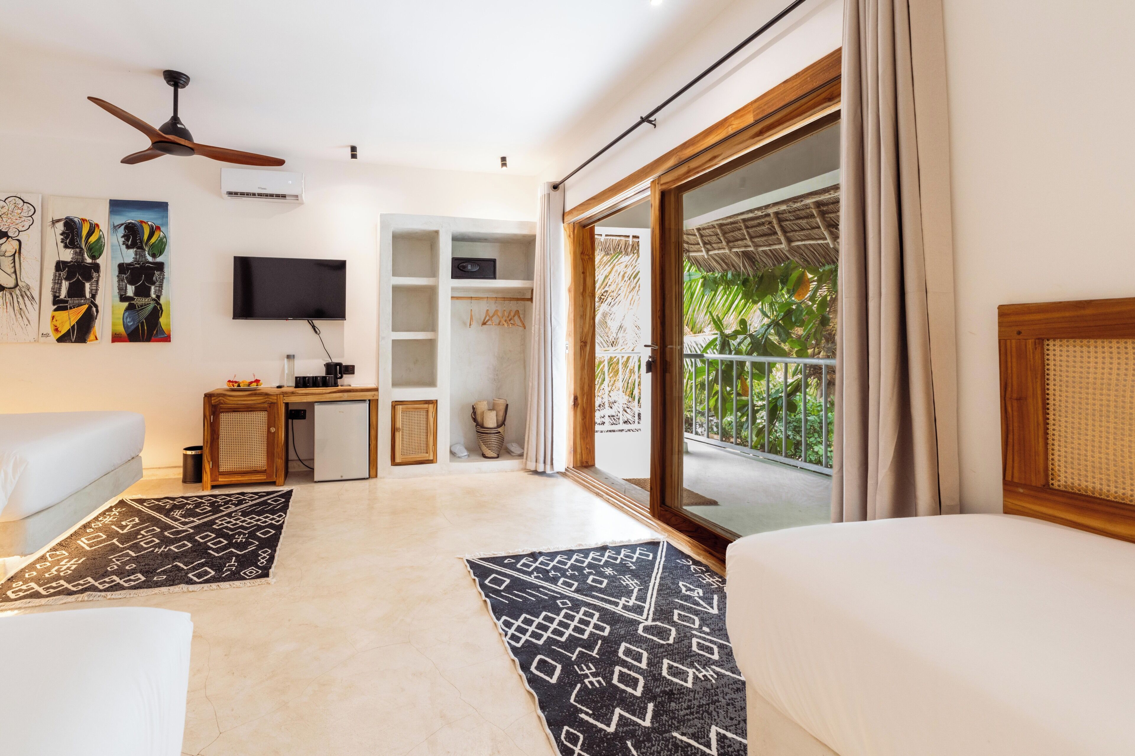 A modern hotel room with a flat-screen TV, ceiling fan, and a balcony with a view of greenery.