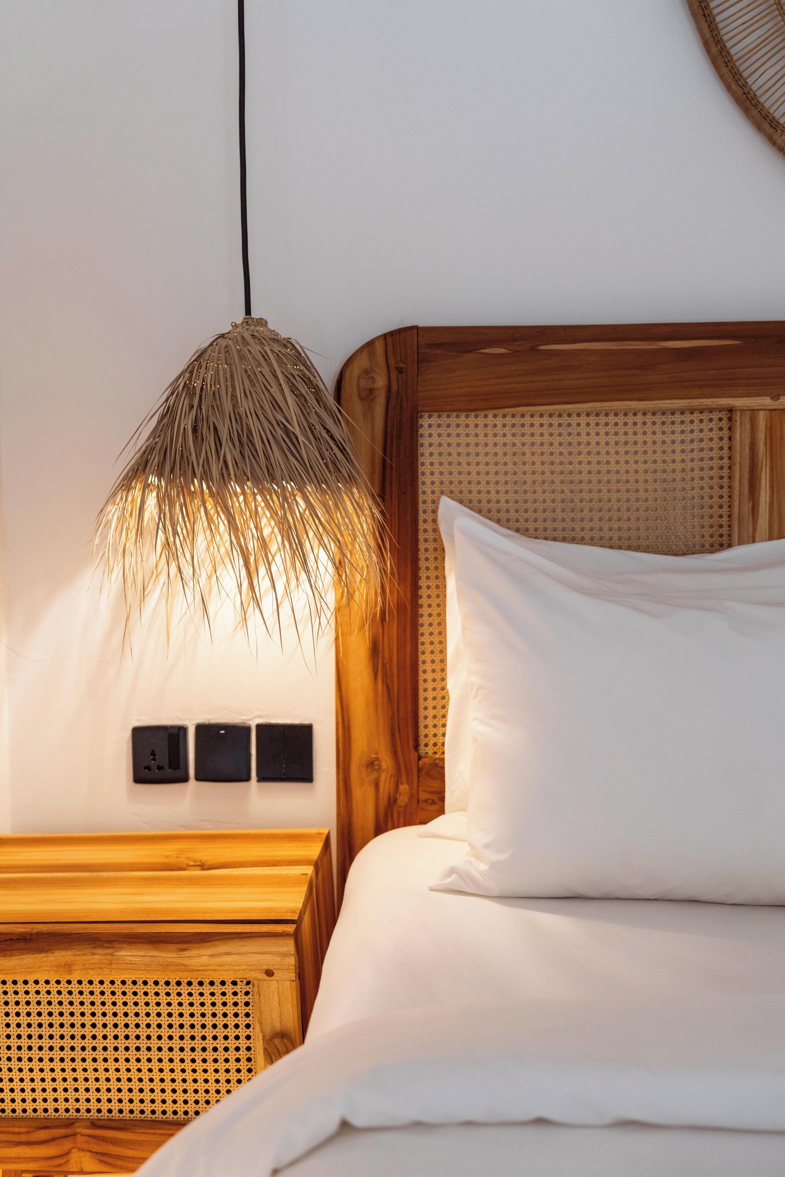 Cama com roupa de cama branca, luminária de fibra natural e cabeceira de madeira.