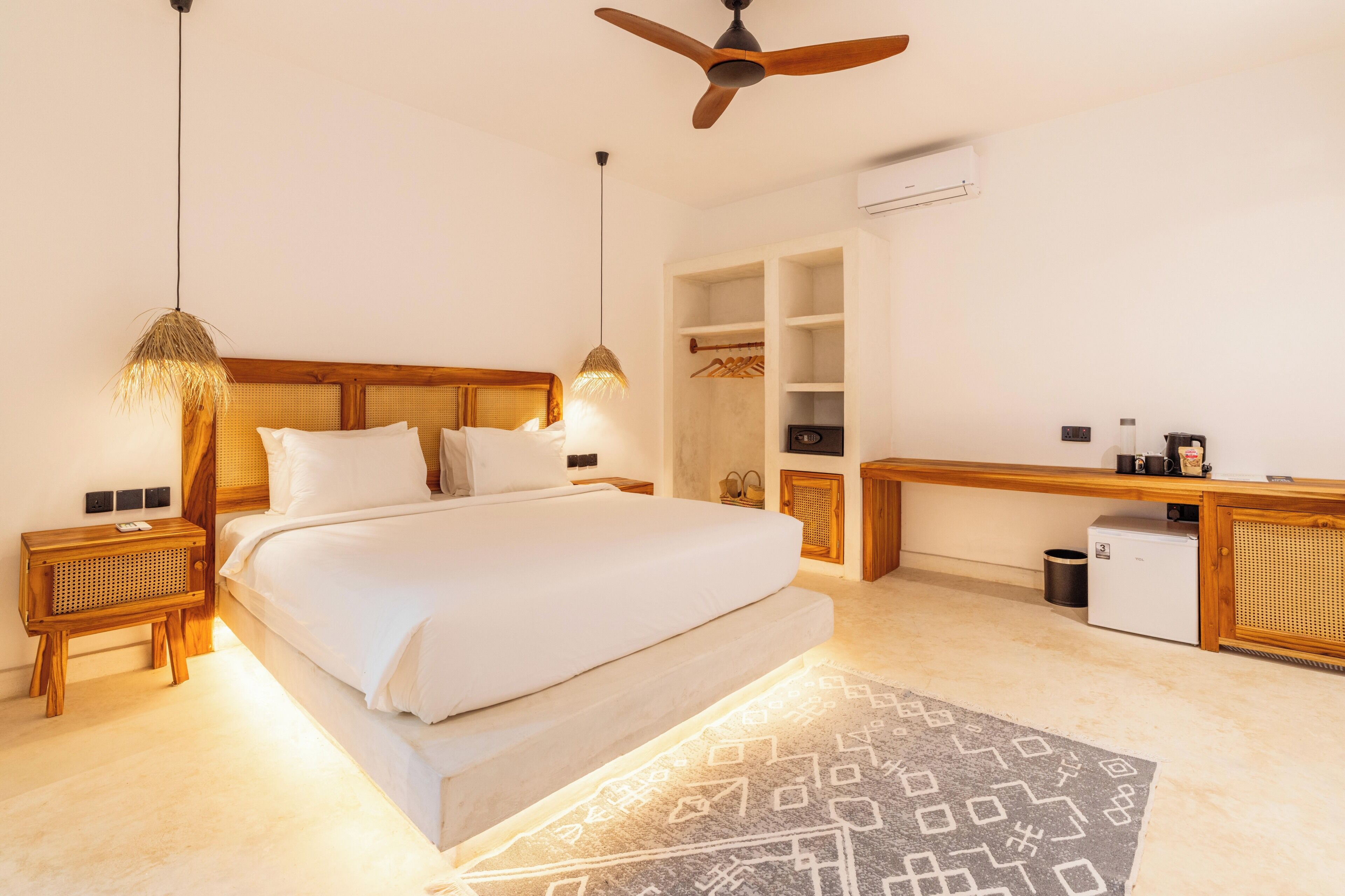 A bedroom with a bed, bedside tables, a ceiling fan, an air conditioning unit, a desk, and a trash bin.