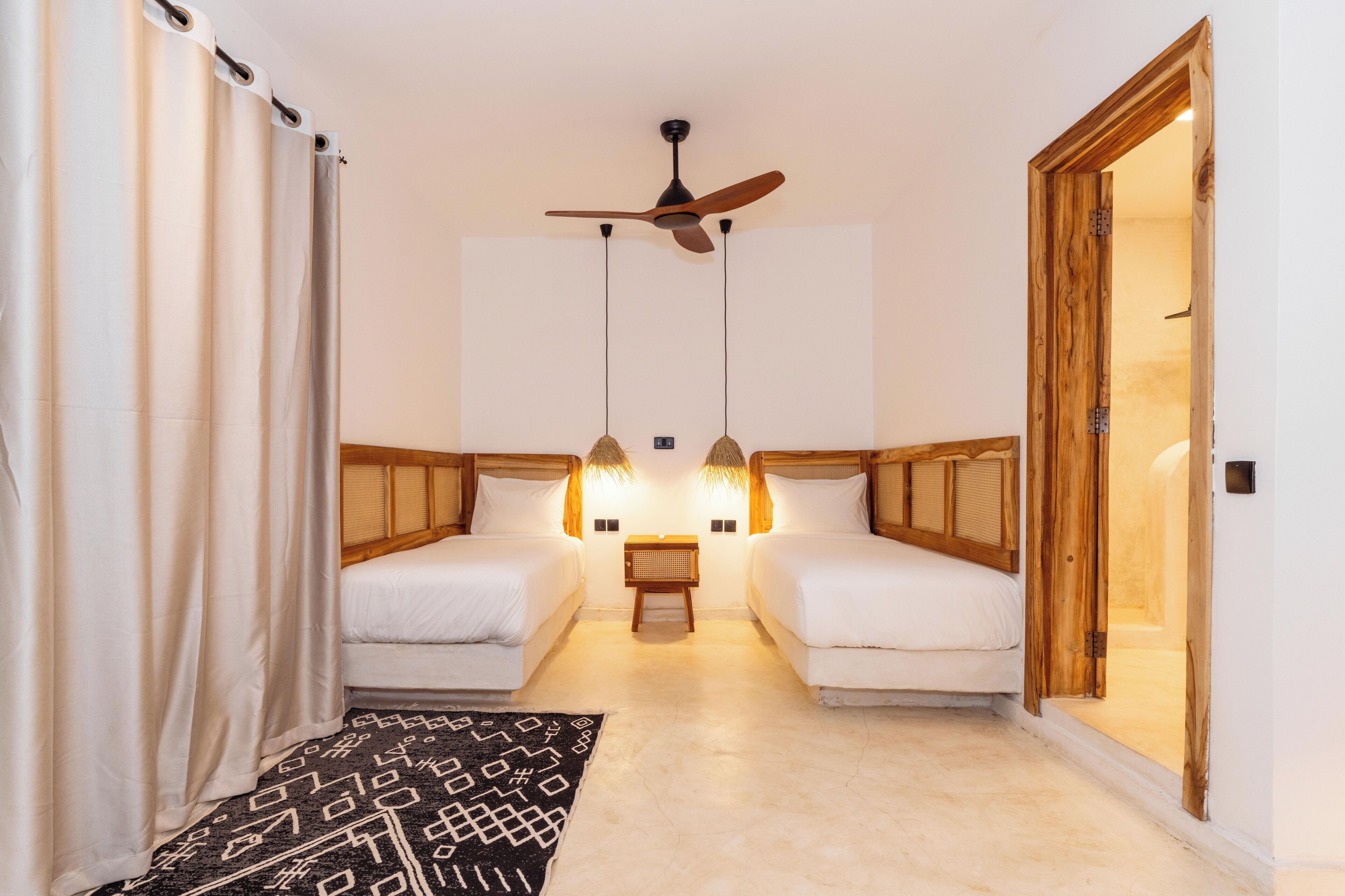 Una habitación con dos camas, ventilador de techo, luces fijadas en la pared, una mesita y una puerta.