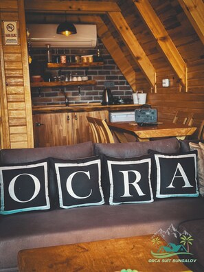 A cozy cabin interior with a wooden dining table, a sofa with 'OCRA' pillows, and a kitchen area with brick walls and wooden beams. - Orca Bungalows (Ardeşen)