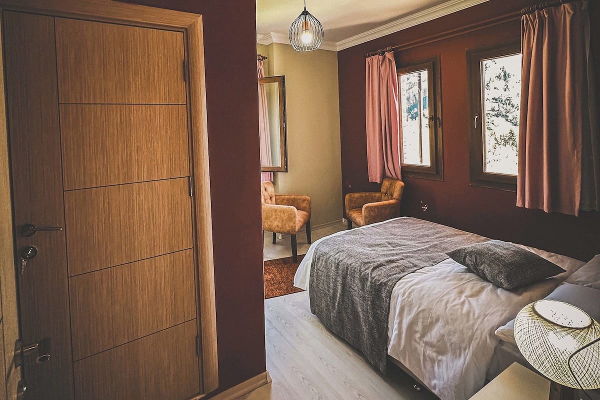 Ein Schlafzimmer mit einem Holzschrank, einem Bett mit weißer und grauer Bettdecke, einer Pendelleuchte und einem Fenster mit Vorhängen.