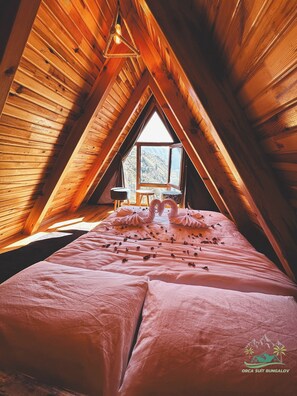 An attic bedroom with a wooden sloped ceiling, a large window, and a bed with pink bedding. - Orca Bungalows (Ardeşen)