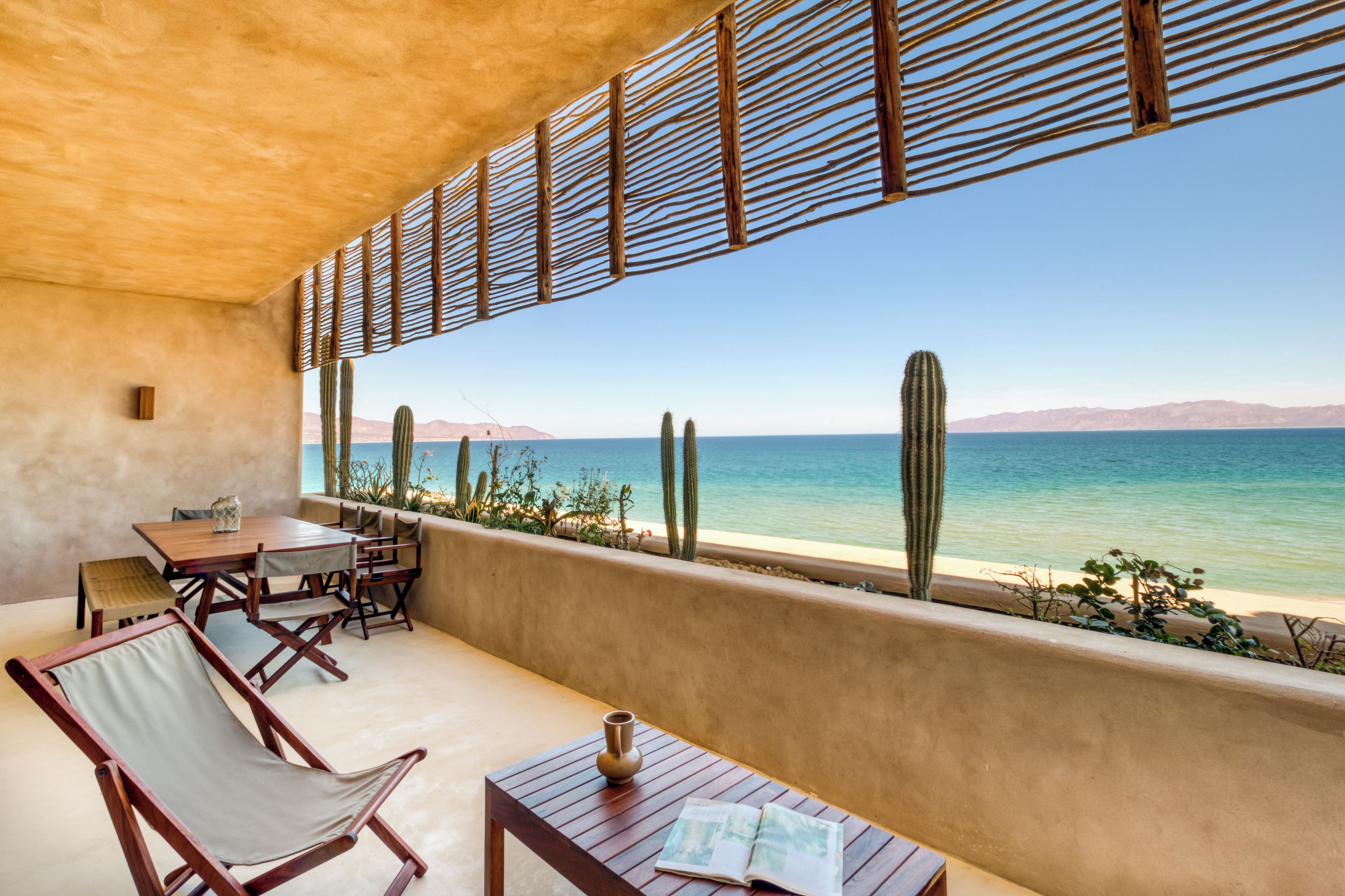 A terrace with a wooden table and chairs, a cactus wall, and a view of the sea.
