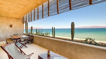 A terrace with a wooden table and chairs, a cactus wall, and a view of the sea.