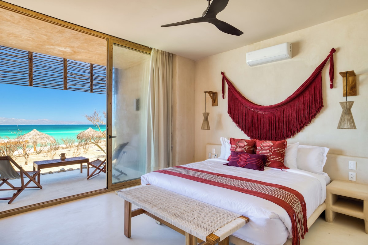 Un dormitorio con una cama grande, una banca, un ventilador de techo y vista a la playa.