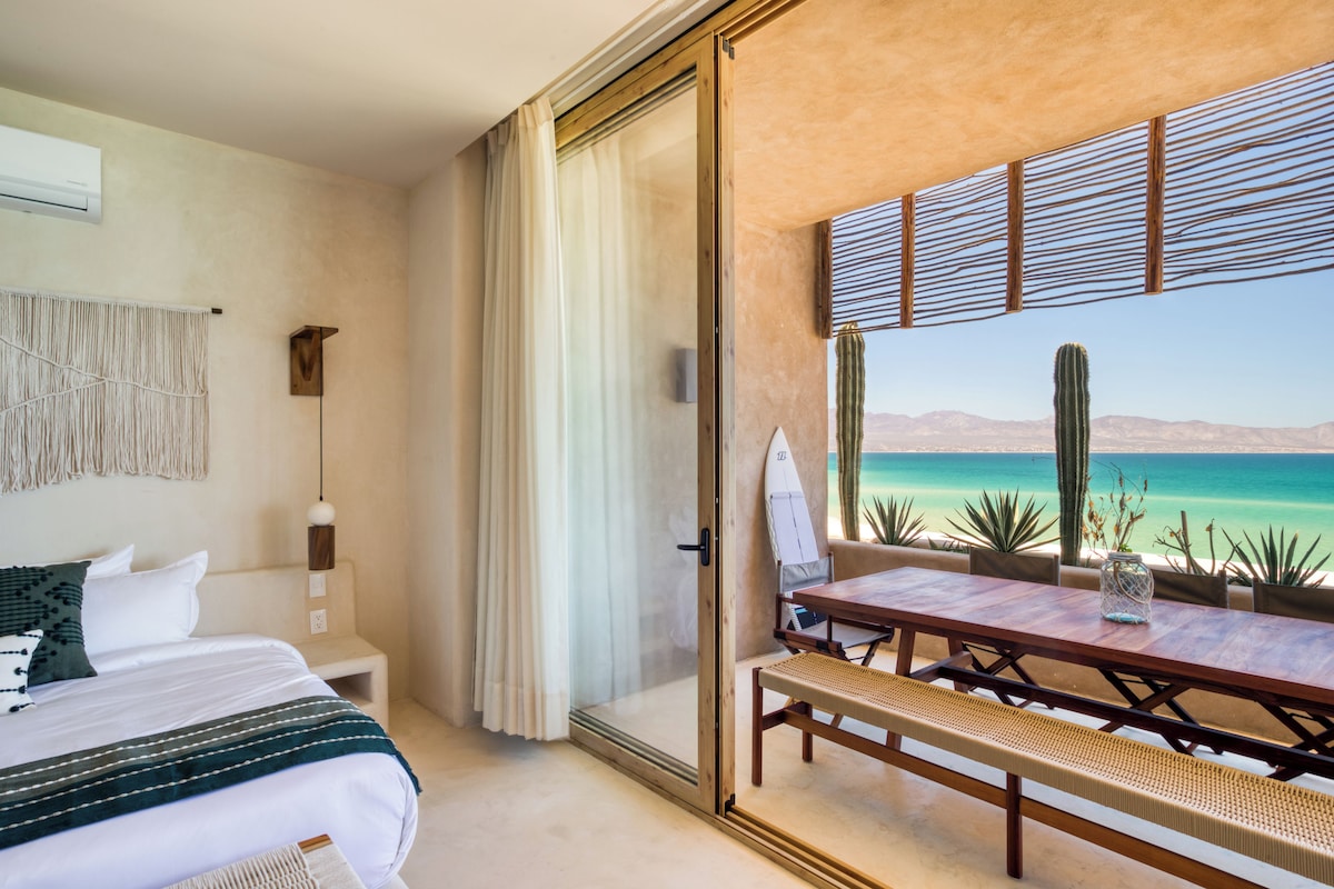 Habitación de hotel con cama, un banco, una mesa y vista al océano.