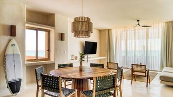 Un salón moderno con una mesa de comedor redonda de madera, rodeada de sillas, un ventanal con cortinas, un televisor montado en la pared y una tabla de surf apoyada contra la pared.