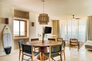 Un salón moderno con una mesa de comedor redonda de madera, rodeada de sillas, un ventanal con cortinas, un televisor montado en la pared y una tabla de surf apoyada contra la pared.