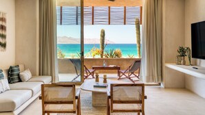 A living room with a sofa, a dining table with chairs, a TV, and a large window with a view of the ocean and mountains. - Amina Wind Resort (La Ventana)