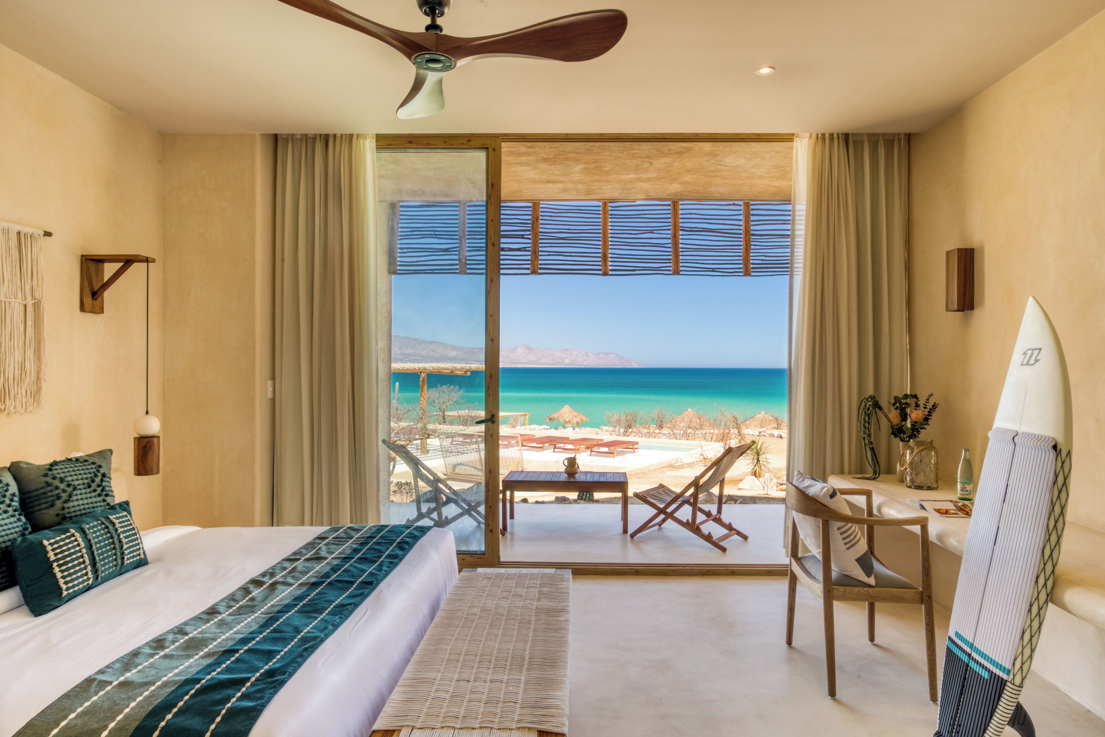 Ein Schlafzimmer mit einem großen Bett, einem Schreibtisch, einem Sessel, einem Surfbrett und Blick auf Strand und Meer.