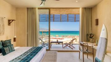 Ein Schlafzimmer mit einem großen Bett, einem Schreibtisch, einem Sessel, einem Surfbrett und Blick auf Strand und Meer.