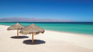 A beach with two straw umbrellas and a mountain range in the background. - Amina Wind Resort (La Ventana)