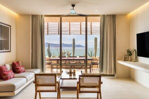 A living room with a sofa, two chairs, a coffee table, a TV, and a large window with a view of the sea and cacti. - Amina Wind Resort (La Ventana)