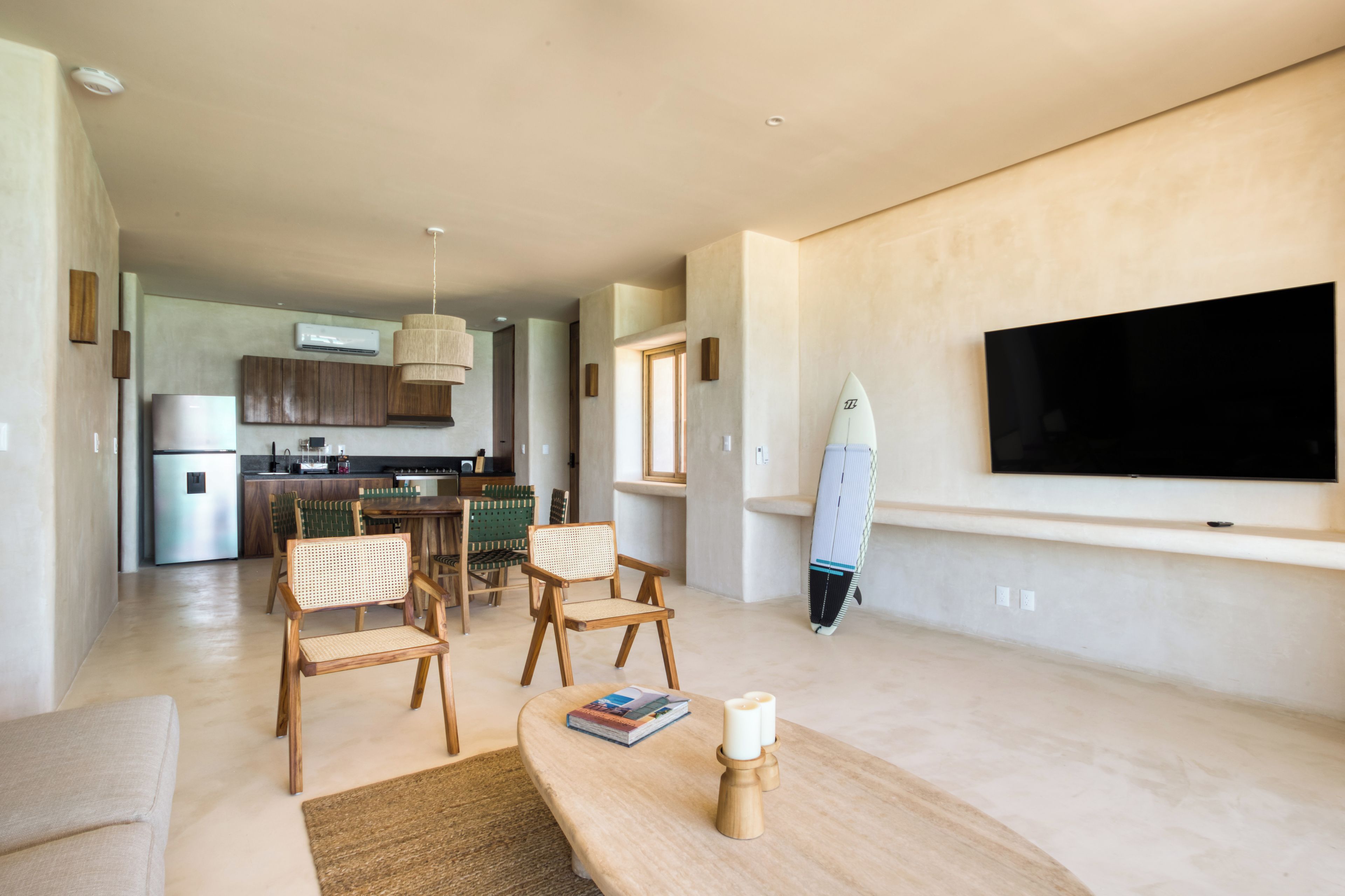 A modern living room with a flat-screen TV, a dining area with wooden chairs, a kitchen in the background, and a surfboard on the wall.