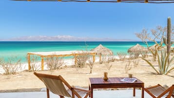 Un área de estar frente a la playa, con mesa y sillas, con vista al mar turquesa y un cielo despejado.