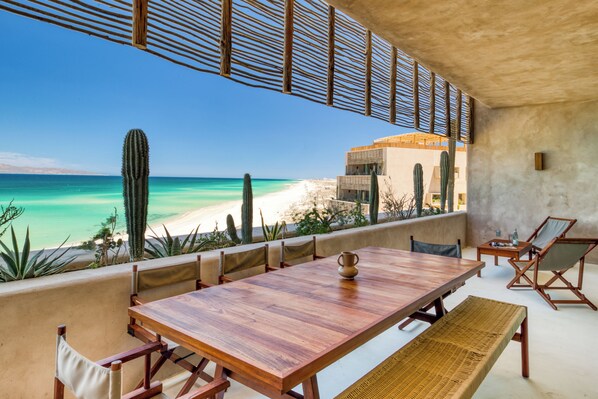A wooden table and chairs set on a balcony overlooking a beach with cacti and a building in the background. - Amina Wind Resort (La Ventana)