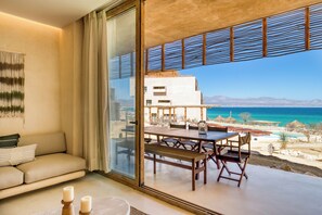 A modern living room with a sofa, a dining table with chairs, and a large glass door leading to a balcony with a view of the sea and mountains. - Amina Wind Resort (La Ventana)