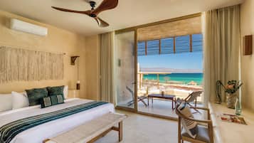 Habitación de hotel con cama, banco, ventilador de techo y balcón con vista a la playa y al océano.