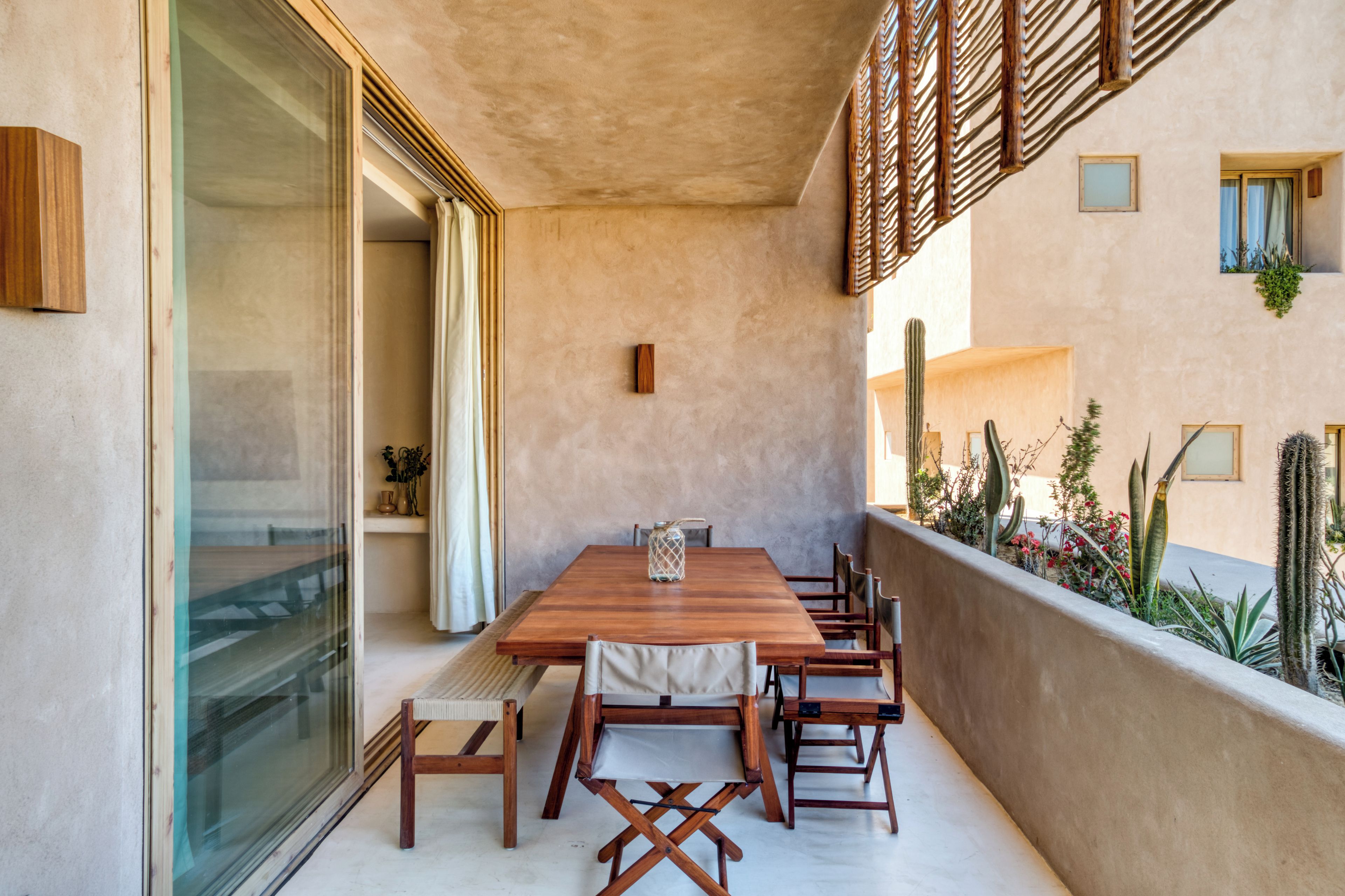 Un balcón con una mesa y sillas de madera, un ventanal de vidrio y un jardín de cactus.